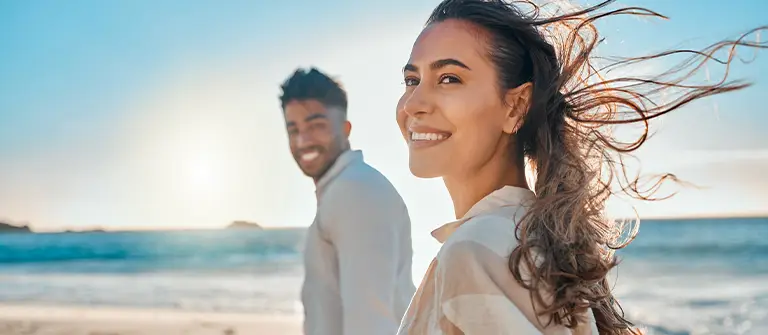 Happy couple smiling on sunny beach with ocean waves and blue sky in background - in Los Angeles, CA