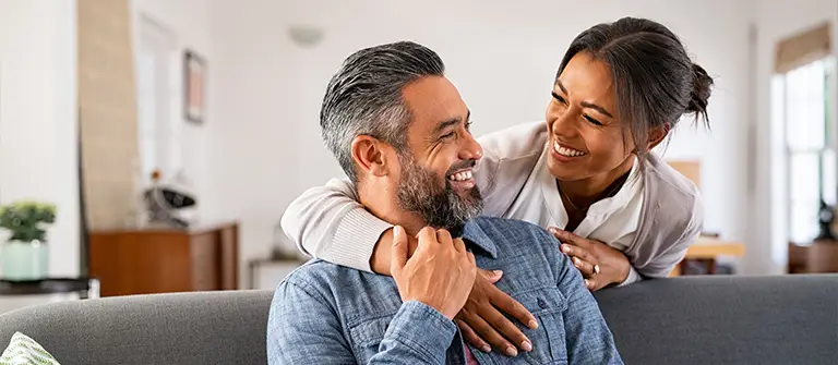 Happy couple embracing on couch at home, woman hugging man from behind while smiling together - Relational in Los Angeles, CA